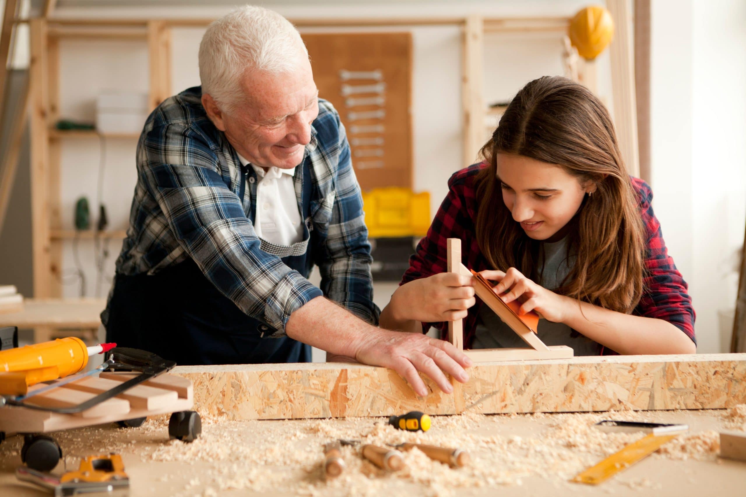An older man and a girl work together on a wood working project in an outdoor building.